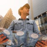 A man stands in front of a cityscape, holding a map and looking thoughtfully at his surroundings