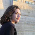 A young woman smiling outdoors near a historical stone wall in Mgarr, Malta.