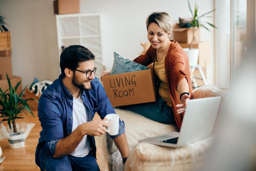 A young couple sits on a couch in their new home, focused on a laptop in front of them