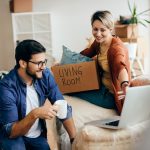 A young couple sits on a couch in their new home, focused on a laptop in front of them